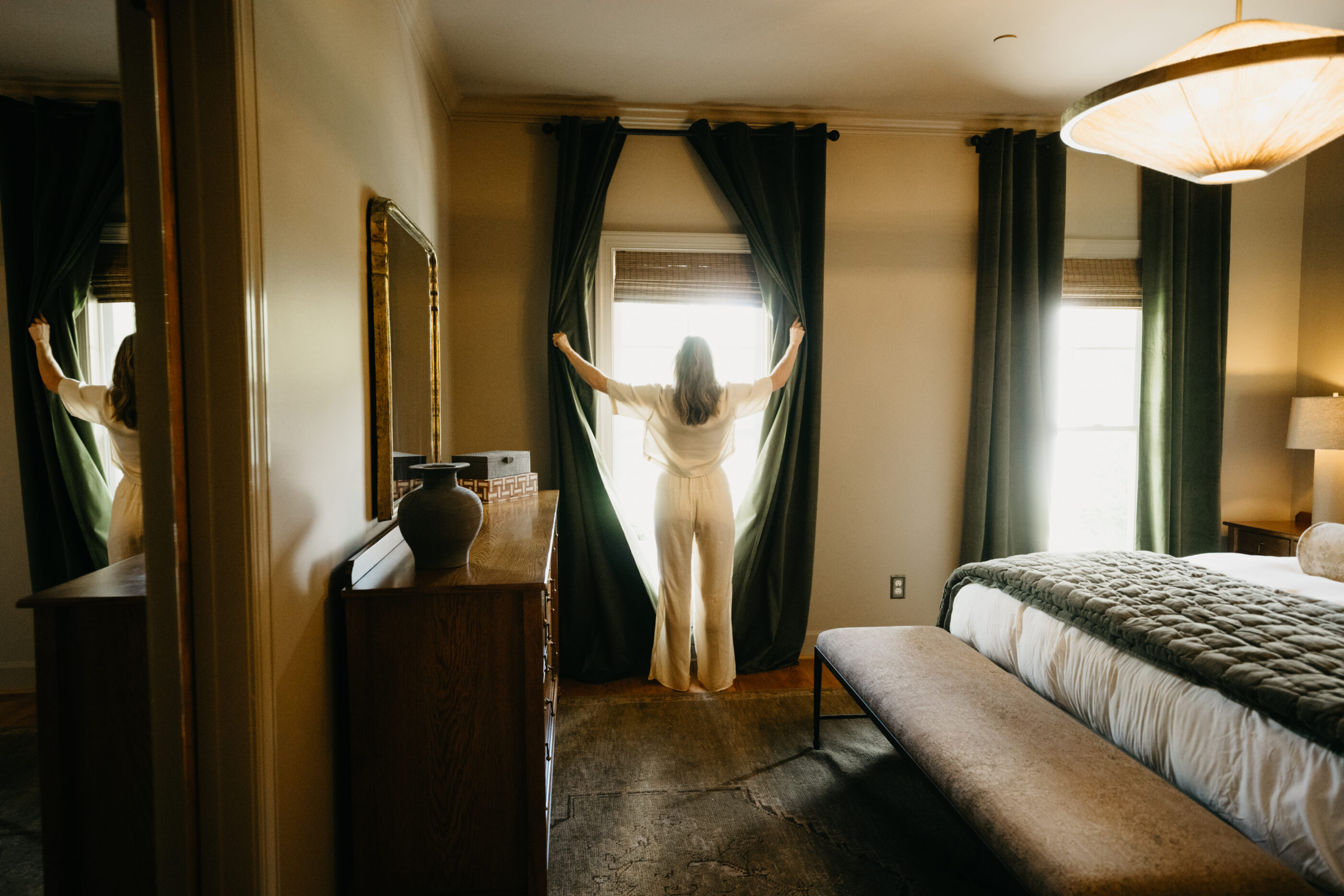 Photo of a woman from behind opening window curtains with sunlight streaming in.